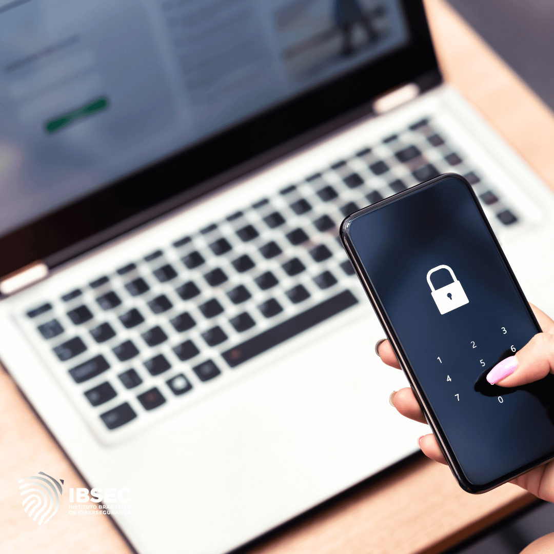 A imagem mostra um laptop aberto em uma mesa, com a tela exibindo uma página de login. Em primeiro plano, uma mão segura um smartphone que também exibe uma tela de bloqueio com um ícone de cadeado. A imagem transmite a ideia de segurança cibernética e autenticação de dois fatores. No canto inferior esquerdo, há um logotipo do IBSEC (Instituto Brasileiro de Segurança).