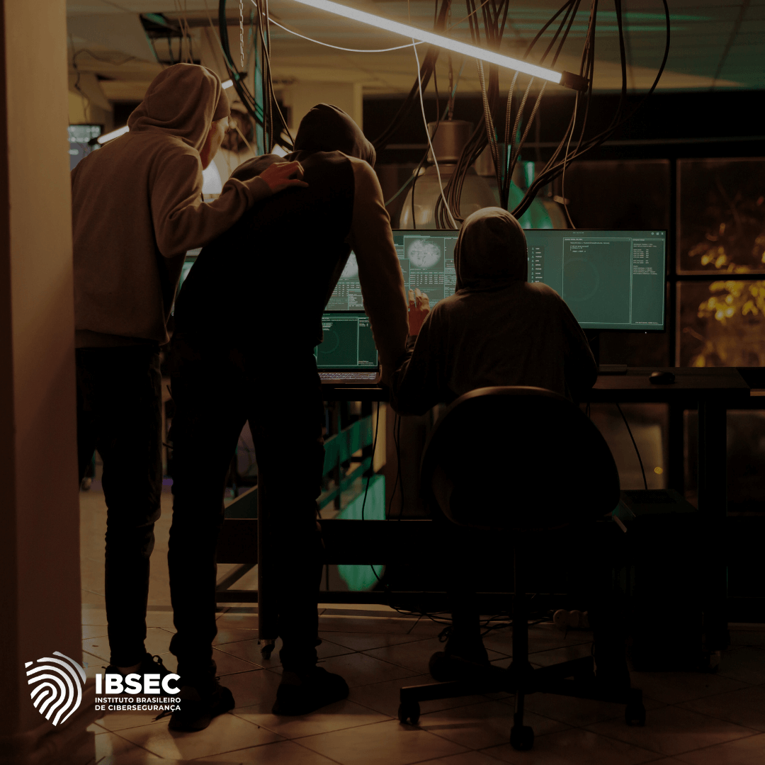 A imagem mostra três pessoas de costas, usando capuzes, reunidas em frente a várias telas de computador em uma sala escura, possivelmente realizando atividades de hacking ou cibersegurança. As telas exibem interfaces de software com gráficos e dados técnicos. O ambiente é cheio de cabos pendurados e iluminação suave, criando uma atmosfera de trabalho técnico e intenso. No canto inferior esquerdo, está o logotipo do IBSEC (Instituto Brasileiro de Cibersegurança).