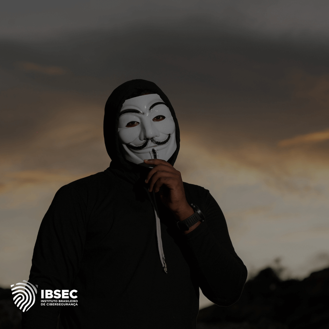 A imagem mostra uma pessoa usando uma máscara de Guy Fawkes, popularmente associada ao grupo hacktivista Anonymous, em um ambiente ao ar livre durante o pôr do sol. A pessoa está vestida com um moletom preto com capuz e mantém um dedo próximo aos lábios, sugerindo silêncio. No canto inferior esquerdo, há o logotipo do IBSEC (Instituto Brasileiro de Cibersegurança).