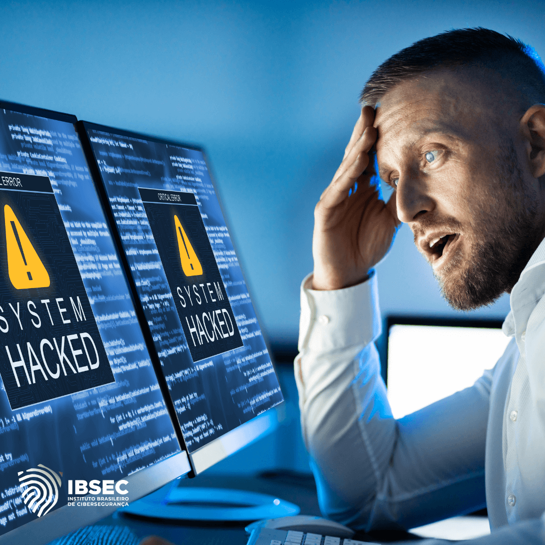 Homem sentado em frente a dois monitores de computador, com expressão de choque e preocupação. Nas telas, aparece a mensagem "SYSTEM HACKED" em destaque, com um ícone de alerta amarelo. No canto inferior esquerdo da imagem, o logotipo do Instituto Brasileiro de Cibersegurança (IBSEC).