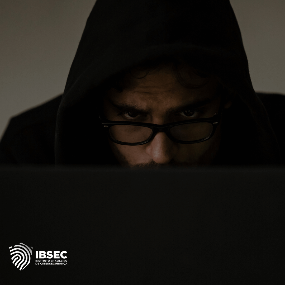 Imagem de uma pessoa usando um capuz preto e óculos, olhando de forma concentrada para um computador em um ambiente escuro. A imagem tem o logotipo do IBSEC - Instituto Brasileiro de Cibersegurança - no canto inferior esquerdo