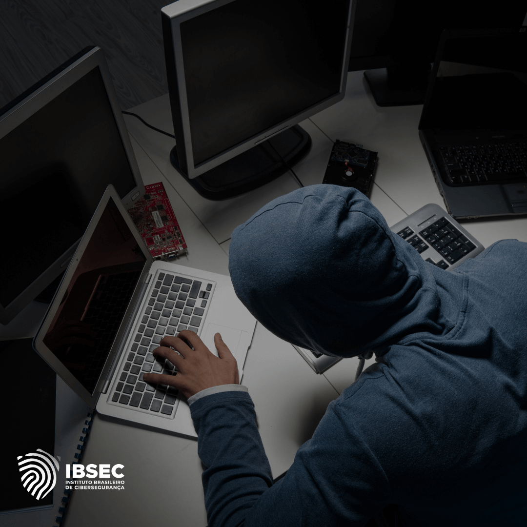 Pessoa com capuz usando um laptop em um ambiente escuro, cercada por vários monitores e equipamentos eletrônicos. A imagem sugere atividades de cibercrime ou hacking. Logotipo do Instituto Brasileiro de Cibersegurança (IBSEC) está localizado no canto inferior esquerdo.