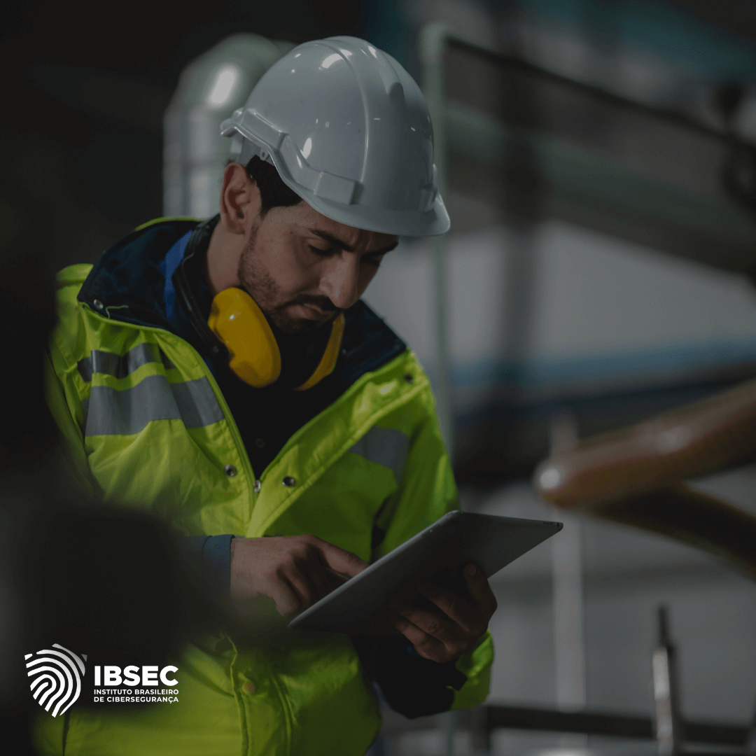 Trabalhador da indústria vestindo equipamento de segurança, incluindo capacete branco e jaqueta refletiva amarela, enquanto utiliza um tablet para realizar verificações em um ambiente industrial. Logotipo do IBSEC no canto inferior esquerdo da imagem.