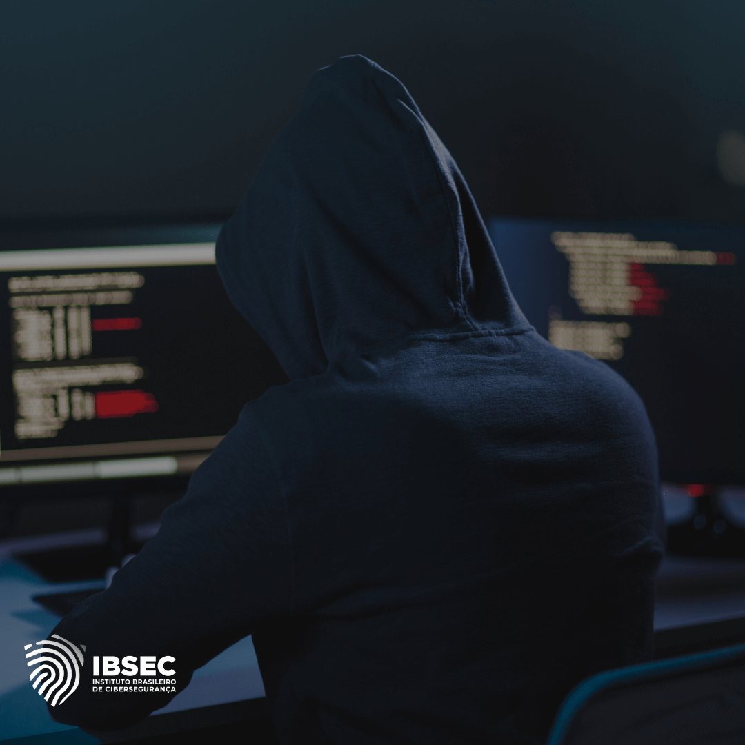 Imagem de uma pessoa com um moletom com capuz, sentada em frente a dois monitores de computador, exibindo códigos. A cena está levemente escurecida, sugerindo um ambiente de hacking ou cibercrime. O logotipo do IBSEC - Instituto Brasileiro de Cibersegurança está presente no canto inferior esquerdo.