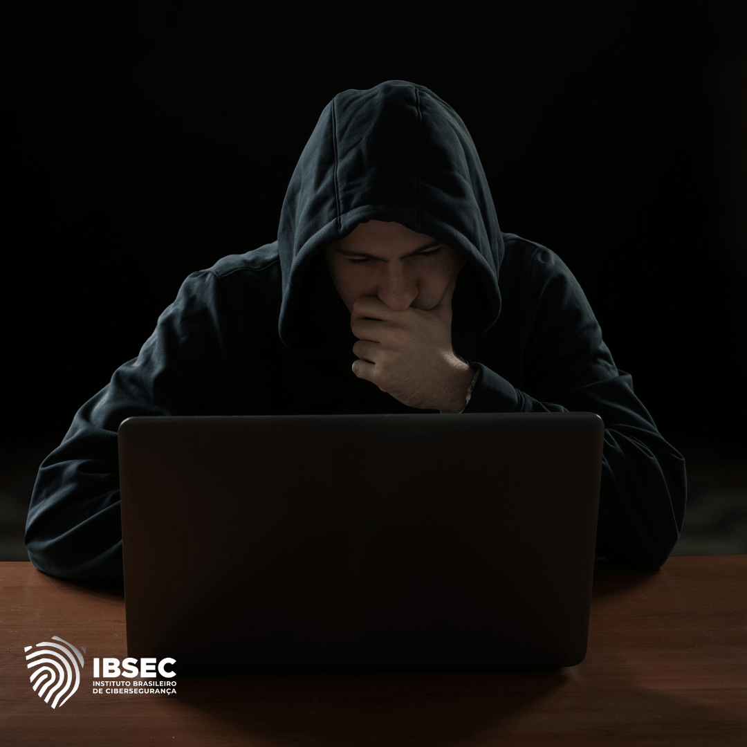 Homem usando moletom com capuz, sentado em frente a um laptop em um ambiente escuro. Ele está concentrado, com a mão no queixo, transmitindo uma imagem de reflexão ou investigação. No canto inferior esquerdo, está o logotipo do IBSEC (Instituto Brasileiro de Cibersegurança).