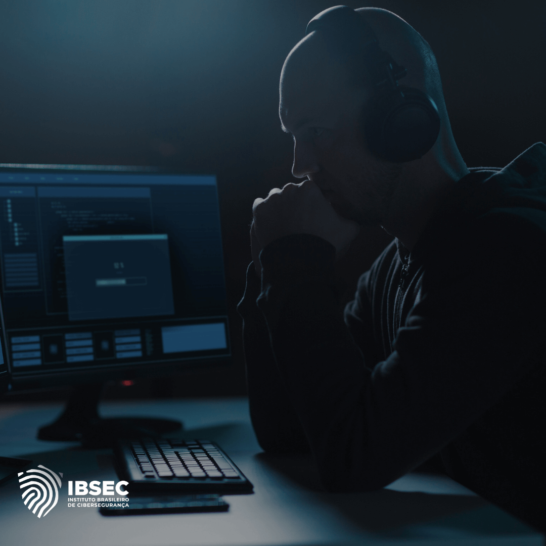 Um homem sentado em frente a dois monitores de computador em um ambiente escuro, usando fones de ouvido. Ele está concentrado, com as mãos próximas ao rosto em uma pose pensativa, enquanto códigos e uma tela de login são visíveis nos monitores. No canto inferior esquerdo, há o logotipo do IBSEC (Instituto Brasileiro de Cibersegurança).