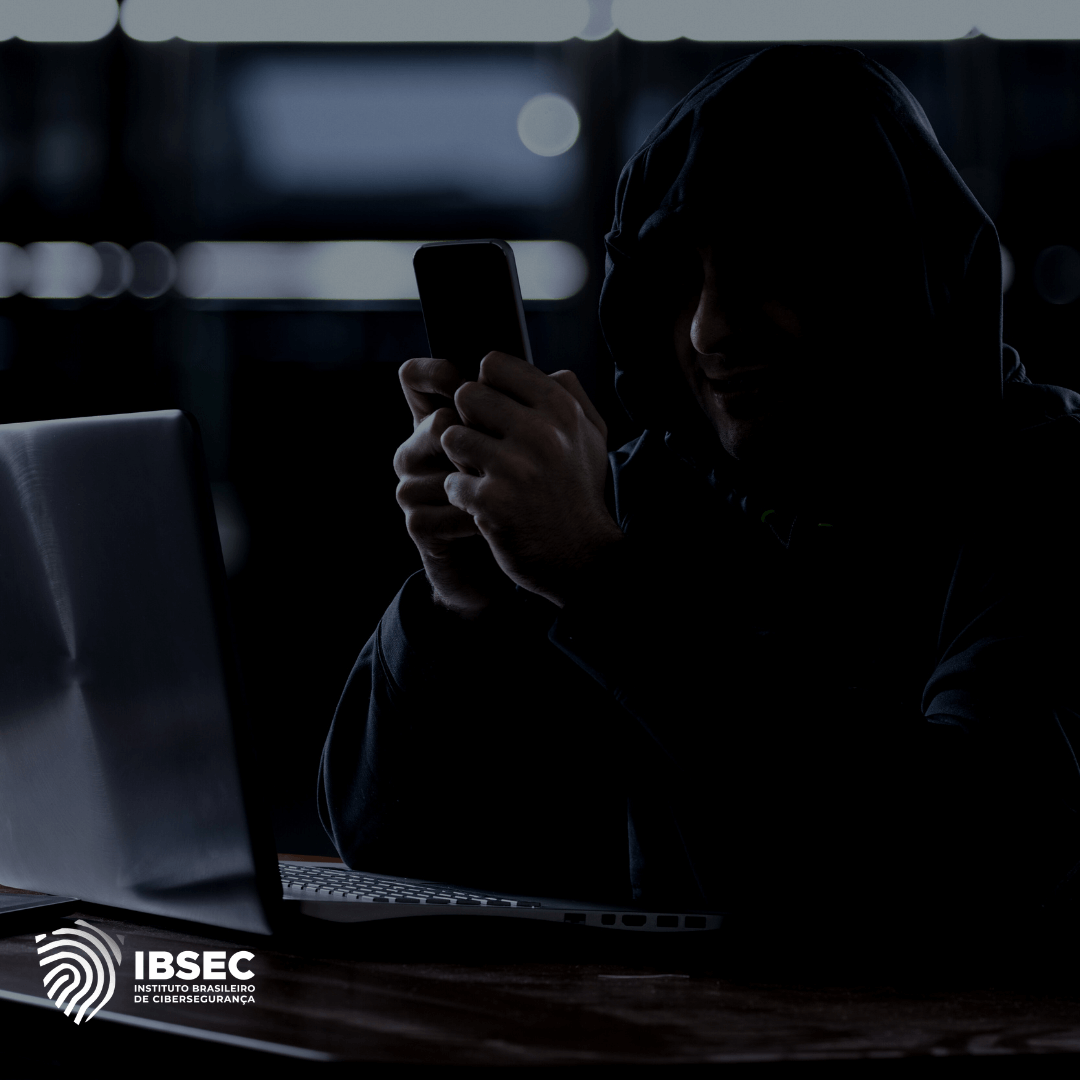 Imagem de um hacker vestido com um moletom com capuz, utilizando um smartphone e trabalhando em um laptop em um ambiente escuro. O logotipo do Instituto Brasileiro de Cibersegurança (IBSEC) está localizado no canto inferior esquerdo.