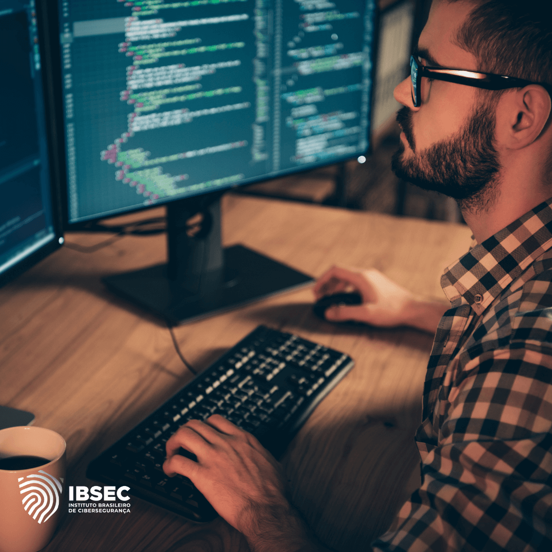 Um homem usando óculos trabalha em um computador de mesa, concentrado em um código que está sendo exibido em dois monitores. Ele usa uma camisa xadrez e está em um ambiente de trabalho com pouca luz. No canto inferior esquerdo da imagem, está o logotipo do IBSEC, Instituto Brasileiro de Cibersegurança. Uma xícara de café está posicionada ao lado do teclado, reforçando o ambiente de concentração e trabalho intenso.