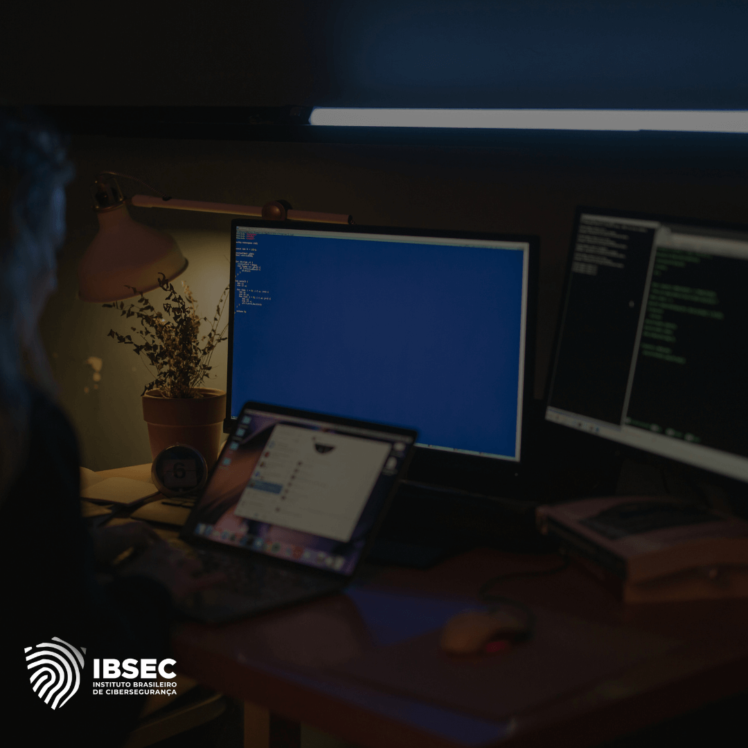 Imagem de uma pessoa trabalhando em um ambiente escuro, com três telas de computador à sua frente. A tela central exibe um fundo azul com linhas de código, enquanto as outras duas mostram interfaces de programação ou scripts em execução. No canto inferior esquerdo, está o logotipo do IBSEC - Instituto Brasileiro de Cibersegurança. O ambiente sugere um trabalho noturno ou em um local de baixa iluminação, enfatizando a seriedade e concentração necessários na área de cibersegurança.