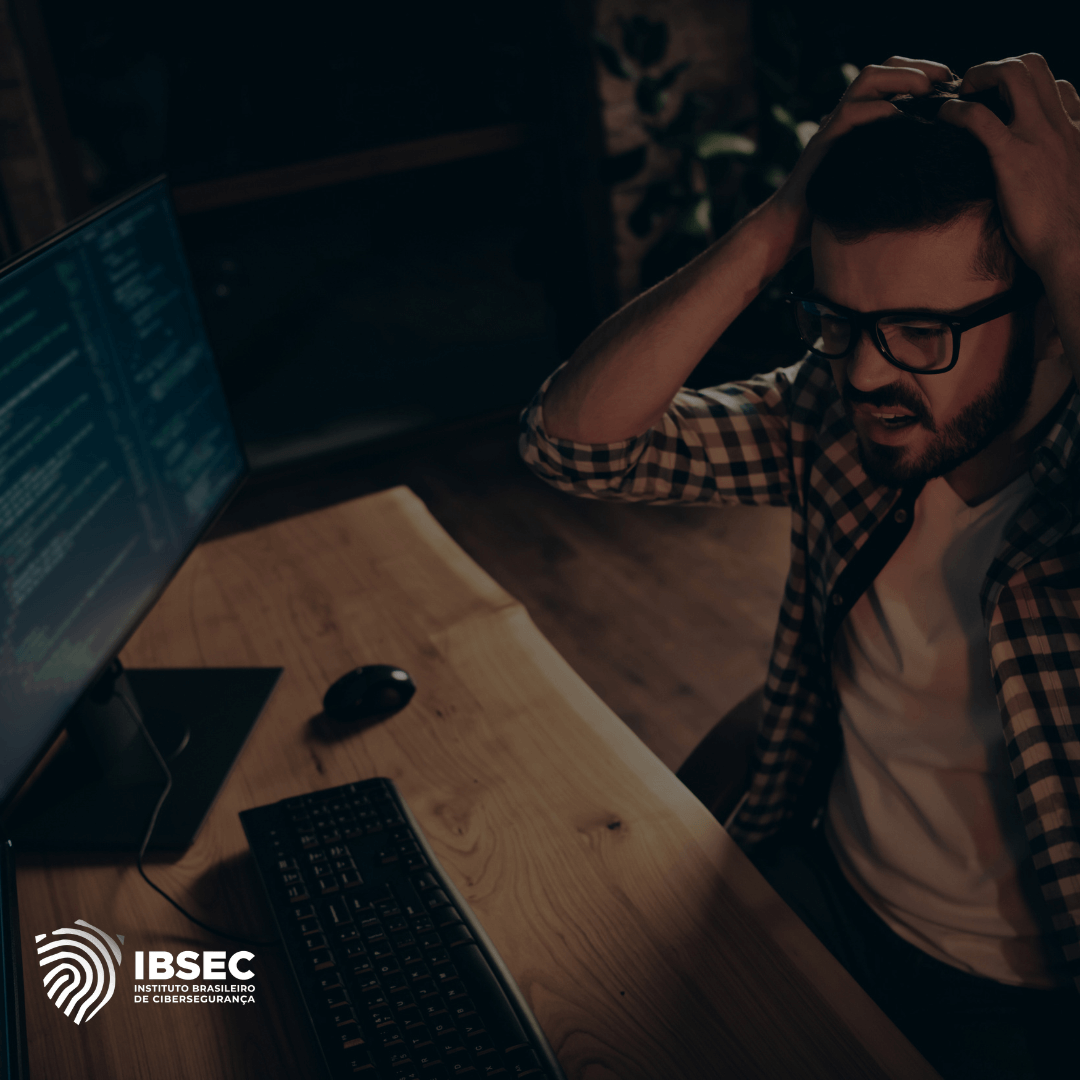 Imagem de um homem sentado à mesa em frente a um computador, demonstrando frustração enquanto segura a cabeça com as mãos. Na tela, há linhas de código, sugerindo um problema técnico ou um possível ataque cibernético. O ambiente parece ser um escritório ou local de trabalho. No canto inferior esquerdo, está o logotipo do IBSEC (Instituto Brasileiro de Cibersegurança), reforçando o tema de cibersegurança e desafios relacionados à proteção de sistemas digitais.