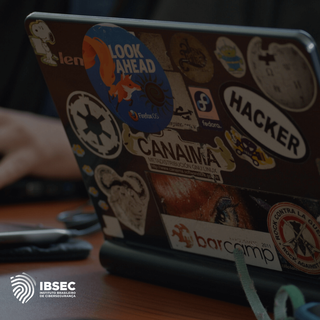 A imagem mostra a parte traseira de um laptop com vários adesivos de temas relacionados à tecnologia, como "Hacker", "Fedora", e "Look Ahead" com a raposa do Firefox OS. Há também adesivos que fazem referência a software livre e cultura hacker, como "Canaima GNU/Linux" e "barcamp". O logo da IBSEC (Instituto Brasileiro de Cibersegurança) aparece no canto inferior esquerdo. A imagem evoca um ambiente tecnológico, voltado para segurança digital e cultura hacker, com ênfase em software de código aberto e inovação.