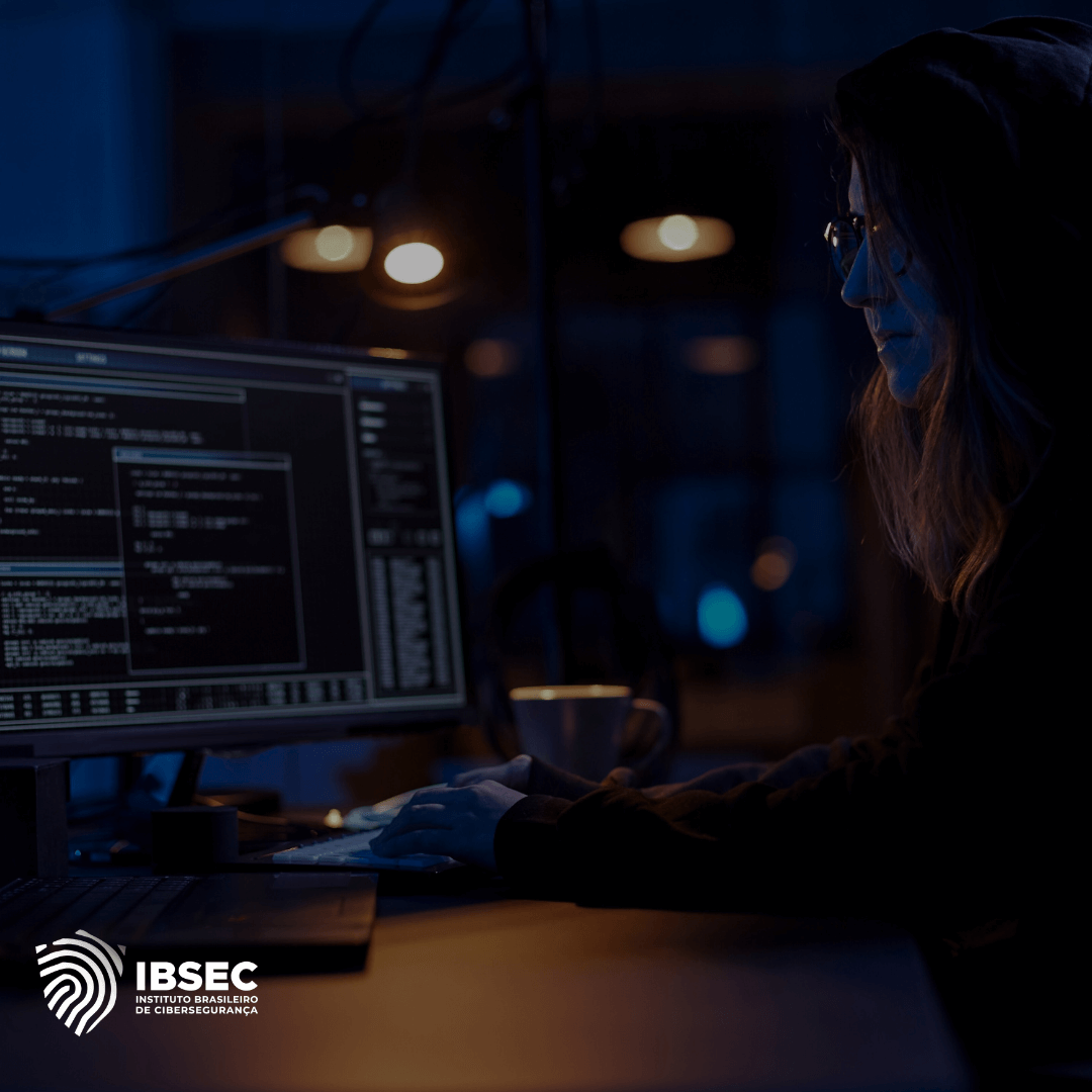 A imagem mostra uma pessoa, possivelmente uma profissional de cibersegurança, trabalhando em um ambiente escuro com pouca iluminação, olhando para uma tela de computador que exibe códigos ou scripts. A atmosfera reflete concentração e vigilância, típicos de atividades como análise de segurança ou detecção de ameaças cibernéticas. No canto inferior esquerdo, aparece o logotipo do "IBSEC - Instituto Brasileiro de Cibersegurança".