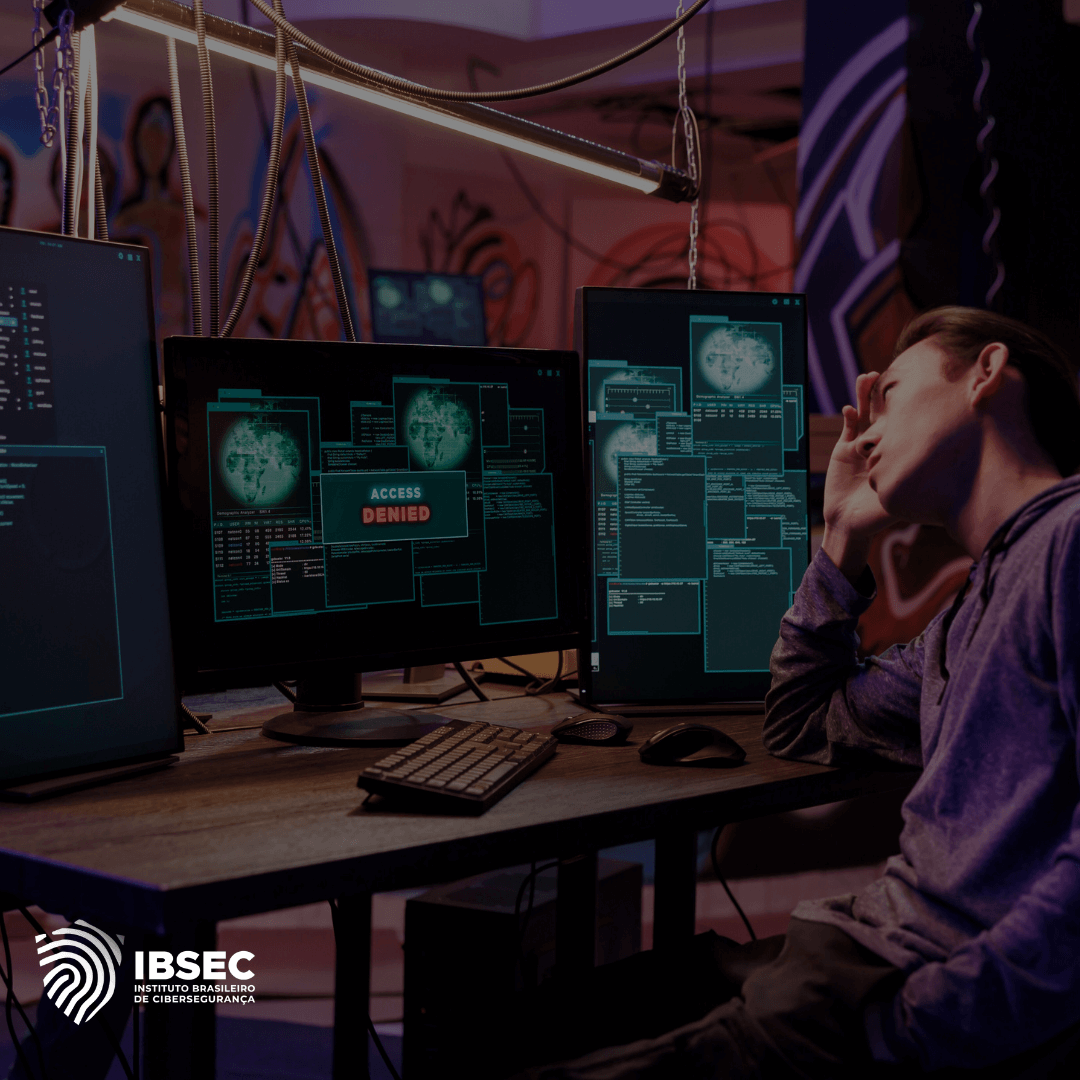 Uma pessoa sentada em frente a vários monitores com sistemas de segurança cibernética, onde uma das telas exibe a mensagem 'Access Denied'. O indivíduo parece frustrado, com a mão na testa. No canto inferior esquerdo, há o logotipo do IBSEC (Instituto Brasileiro de Cibersegurança)