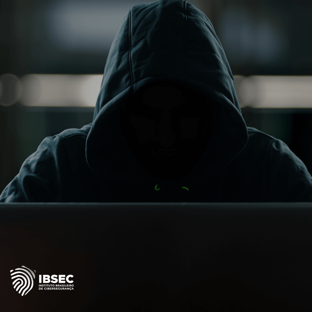 Imagem de um hacker em um ambiente escuro, com capuz cobrindo parcialmente o rosto, olhando para a tela de um laptop. O logo do Instituto Brasileiro de Cibersegurança (IBSEC) está visível no canto inferior esquerdo. A imagem transmite a ideia de ameaça cibernética ou ataque hacker.