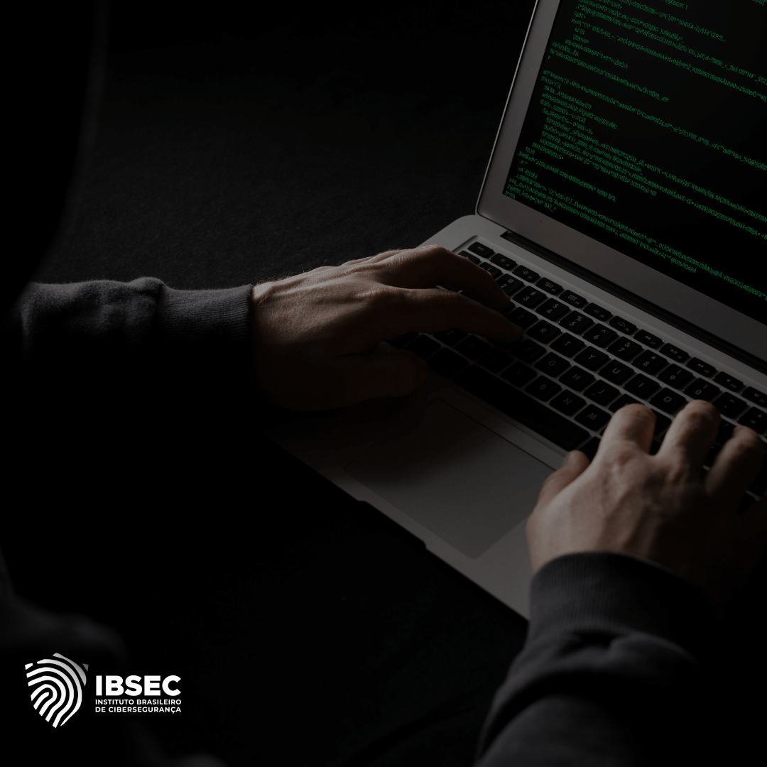 Imagem de uma pessoa digitando em um laptop com código em verde exibido na tela, sugerindo atividade de programação ou possível ataque cibernético. A cena está em um ambiente escuro, reforçando o clima de sigilo e ameaça. O logo do Instituto Brasileiro de Cibersegurança (IBSEC) está visível no canto inferior esquerdo. A imagem transmite a ideia de segurança cibernética ou invasão hacker.
