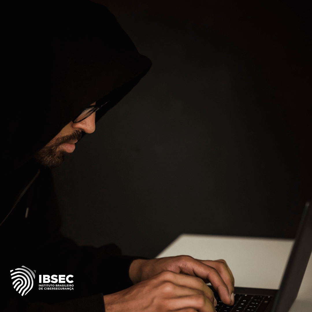 Imagem de um homem usando capuz e óculos, concentrado enquanto digita em um laptop em um ambiente escuro. A cena sugere uma atividade de hacking ou cibercrime, enfatizando o clima de sigilo e ameaça. No canto inferior esquerdo, está o logotipo do IBSEC (Instituto Brasileiro de Cibersegurança).