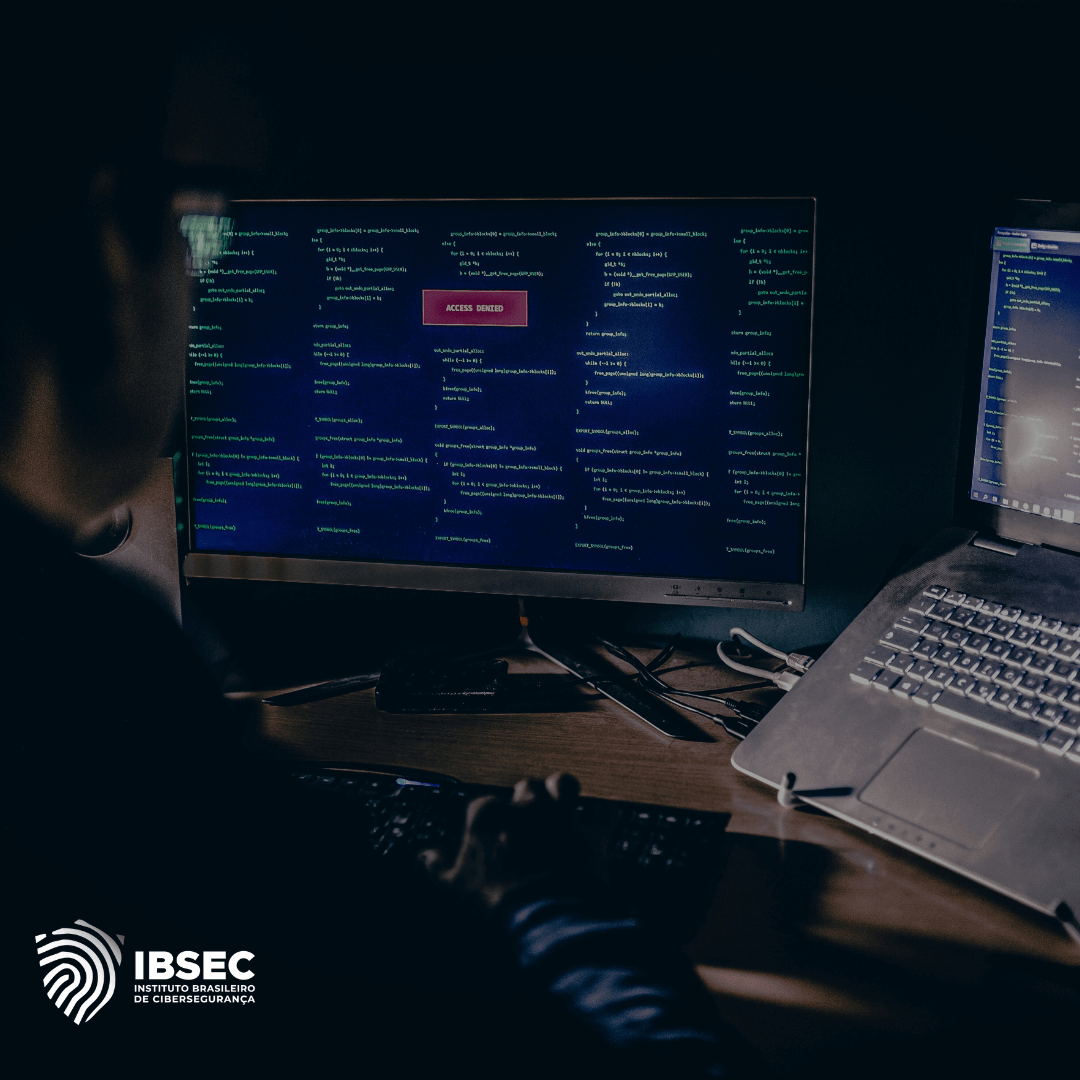 A imagem mostra uma pessoa em um ambiente de baixa iluminação sentada em frente a dois computadores. Um dos monitores exibe linhas de código ou comandos, com um alerta em destaque escrito "Access Denied" no centro da tela. O indivíduo parece estar envolvido em uma atividade cibernética, potencialmente relacionada a segurança da informação ou tentativas de violação. O logo no canto inferior esquerdo pertence ao Instituto Brasileiro de Cibersegurança (IBSEC). O cenário sugere um ambiente de trabalho em cibersegurança ou uma simulação de ataque hacker.