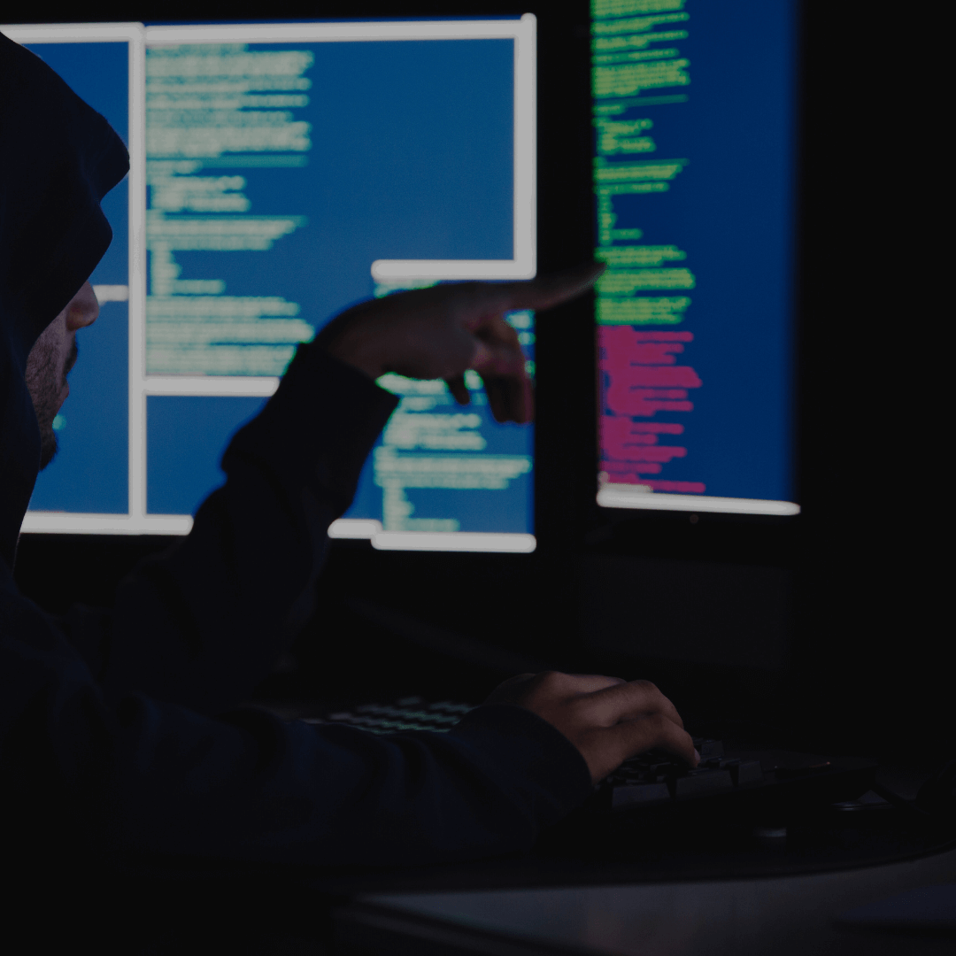 Um indivíduo usando um moletom com capuz, apontando para um monitor com códigos e scripts, em um ambiente escuro. A tela exibe várias janelas com códigos em fundo azul e verde, sugerindo uma análise de dados ou atividade de programação em um ambiente de cibersegurança.