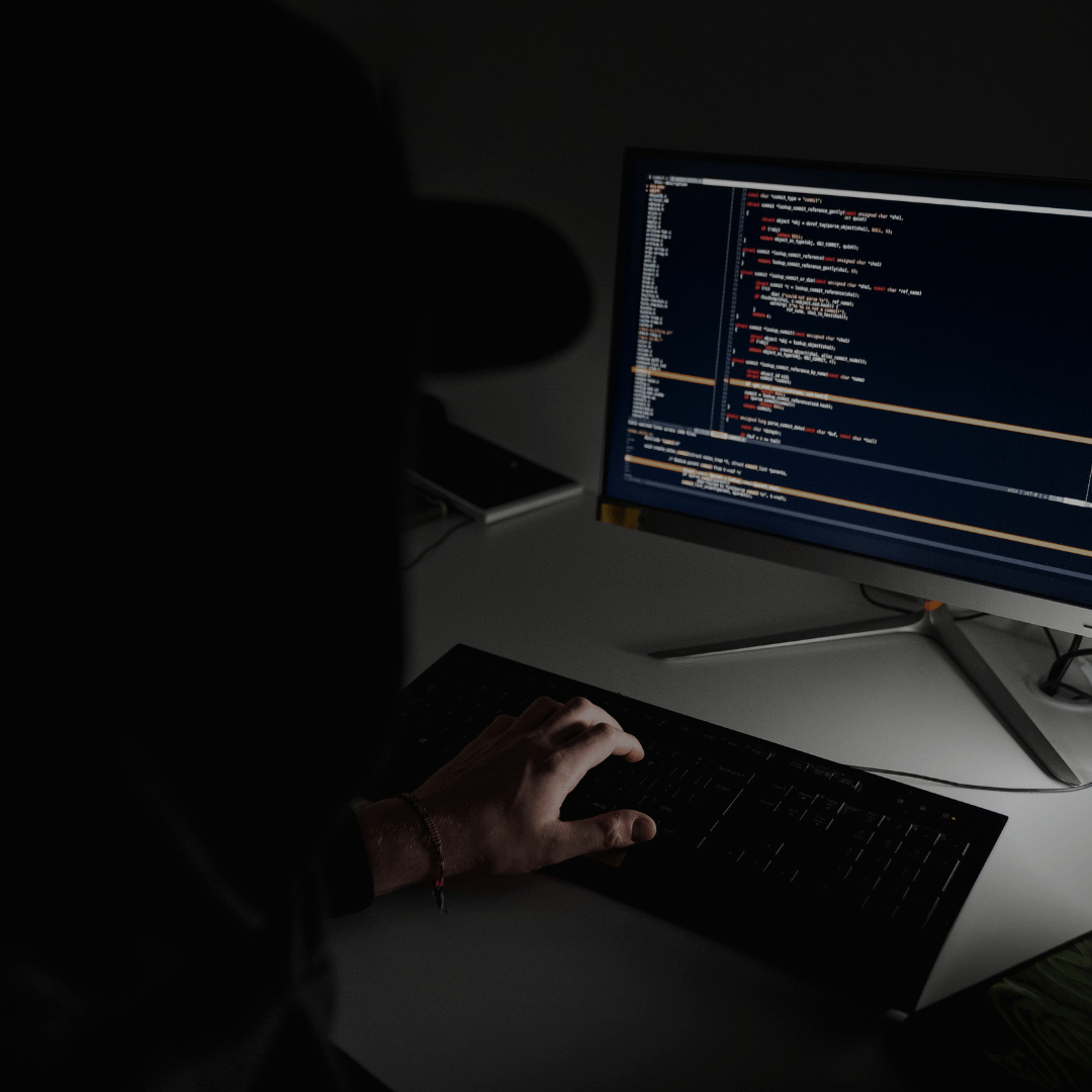 Uma pessoa em um ambiente escuro usando um computador de mesa, com um monitor que exibe linhas de código. A imagem é focada na mão da pessoa digitando no teclado, sugerindo um ambiente de programação, desenvolvimento de software ou possivelmente atividades de hacking. A iluminação baixa e a pose da pessoa criam uma atmosfera de sigilo e mistério.