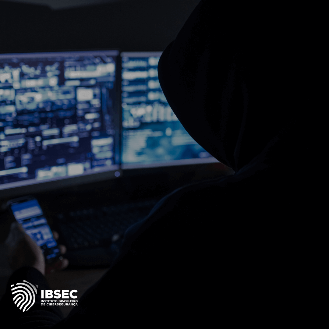 Pessoa com capuz visualizada de costas, segurando um smartphone enquanto trabalha em frente a dois monitores que exibem várias informações digitais e gráficos, sugerindo uma atividade de monitoramento ou invasão cibernética. O ambiente é escuro, reforçando o clima de sigilo e cibersegurança. No canto inferior esquerdo, está o logotipo do IBSEC - Instituto Brasileiro de Cibersegurança.