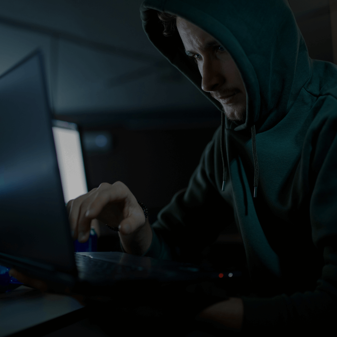 Homem usando um moletom com capuz, operando um laptop em um ambiente escuro. A expressão facial séria sugere concentração, e a iluminação baixa cria um clima de sigilo, associado frequentemente à ideia de atividades cibernéticas como hacking.