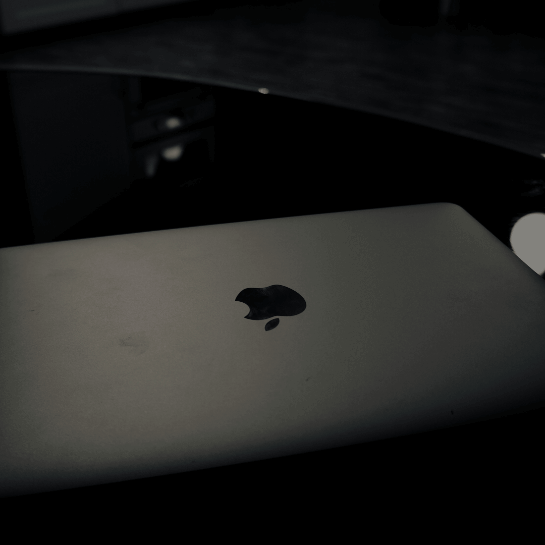 Imagem de um laptop fechado com o logotipo da Apple em destaque, sobre uma superfície escura. O ambiente ao redor é pouco iluminado, criando um contraste entre o brilho suave do logotipo e a escuridão do fundo. A cena transmite uma sensação de minimalismo e foco, sugerindo um espaço de trabalho ou uma área onde dispositivos tecnológicos são usados discretamente.