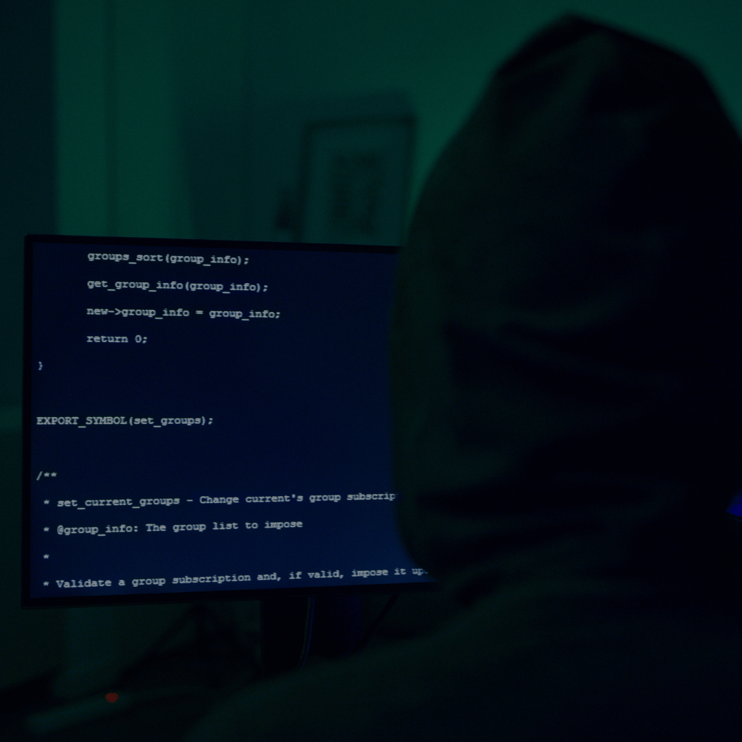 Imagem de uma pessoa usando um capuz, sentada em frente a um monitor de computador em um ambiente escuro. Na tela, é possível ver código de programação exibido, sugerindo atividades de desenvolvimento ou possivelmente hacking. A iluminação baixa e a cor esverdeada da luz criam uma atmosfera misteriosa e clandestina, frequentemente associada ao mundo dos cibercriminosos ou à programação avançada. A identidade da pessoa não é revelada, reforçando o tom anônimo e secreto da cena.