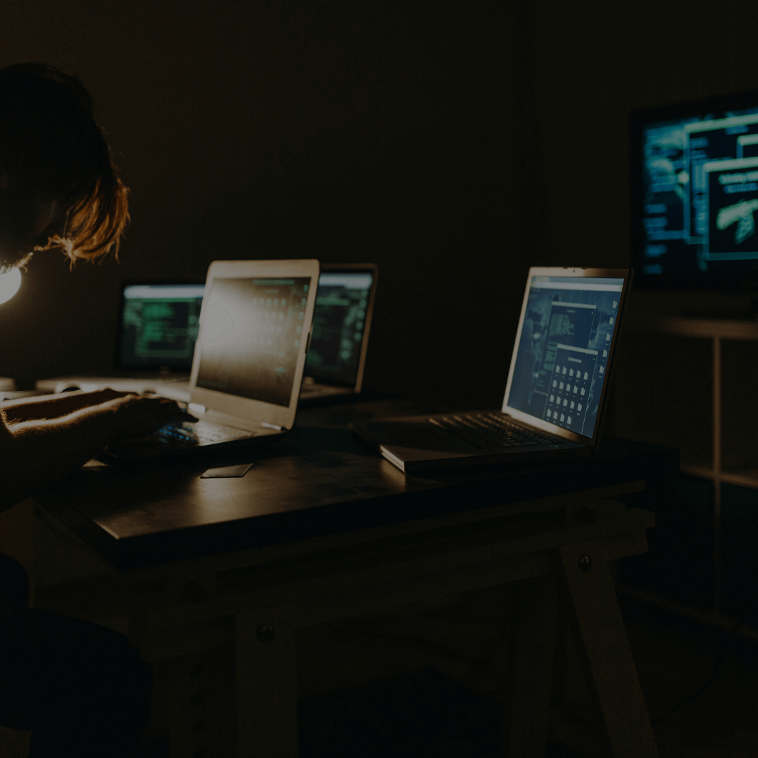 Uma pessoa trabalhando em um ambiente de pouca luz, cercada por vários laptops com telas exibindo códigos e interfaces digitais. No fundo, há uma grande tela mostrando gráficos e informações de segurança cibernética, sugerindo uma operação ou monitoramento relacionado a tecnologia e segurança da informação.