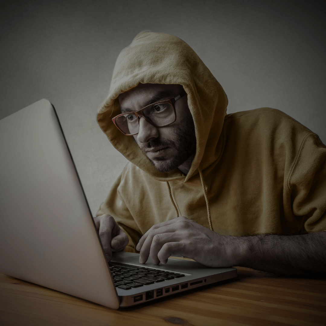 Homem usando moletom com capuz e óculos, concentrado em um laptop. A imagem sugere uma atividade digital ou cibernética, comumente associada a um hacker ou alguém envolvido em atividades online sigilosas.