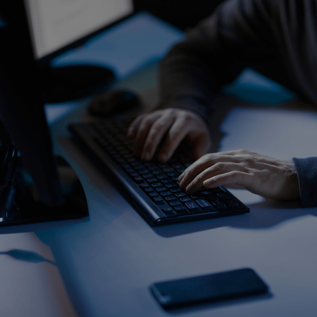 Pessoa digitando em um teclado de computador em um ambiente escuro, com um smartphone sobre a mesa ao lado. A cena sugere atividades relacionadas a tecnologia, possivelmente envolvendo cibersegurança ou trabalho noturno.