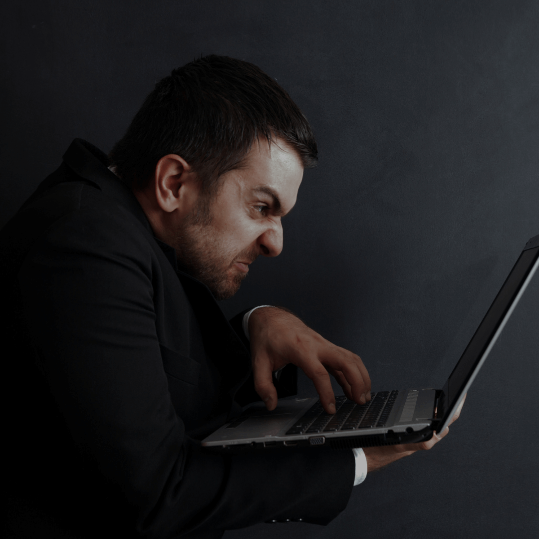 Homem em traje social usando um laptop com expressão facial de frustração e raiva, inclinado para a frente e pressionando as teclas com força. A imagem sugere estresse ou irritação enquanto ele trabalha no computador, com fundo escuro e atmosfera tensa.