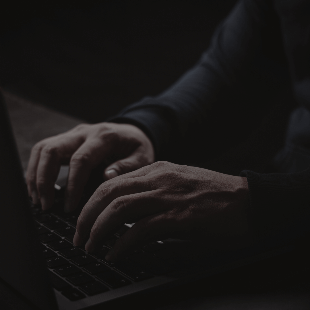 Mãos digitando em um teclado de computador, em um ambiente com pouca iluminação, sugerindo uma atividade digital ou cibersegurança.