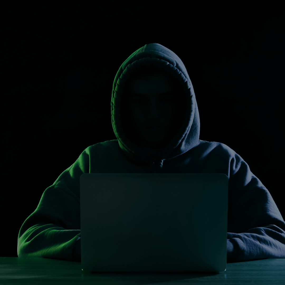 Pessoa vestindo um moletom com capuz sentada em frente a um laptop em um ambiente escuro, com iluminação suave, sugerindo um cenário de atividades cibernéticas ou hacking