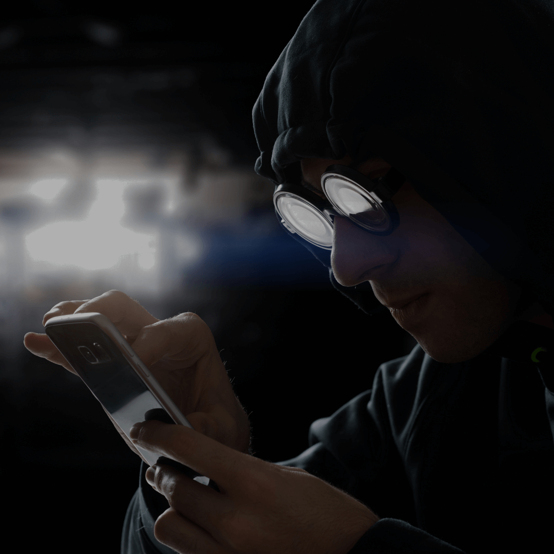 Imagem de uma pessoa usando um capuz e óculos de proteção grandes, olhando atentamente para um smartphone. O ambiente é escuro e cria uma atmosfera misteriosa e sombria, sugerindo uma atividade clandestina ou suspeita, como hacking ou espionagem digital. A expressão concentrada da pessoa e o uso do capuz e dos óculos reforçam o estereótipo de um hacker, trabalhando discretamente em um local pouco iluminado.