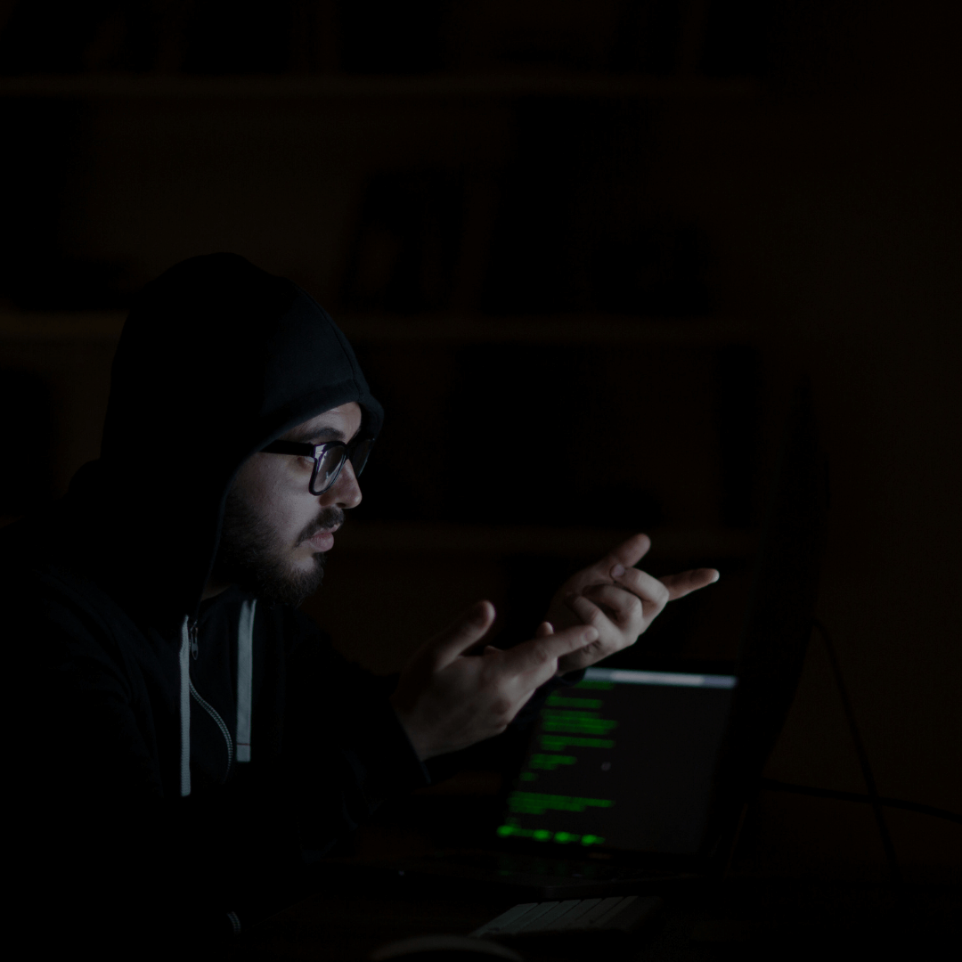 Pessoa em ambiente escuro, usando capuz e óculos, gesticulando em frente a um laptop com código verde na tela, representando um possível hacker ou profissional de cibersegurança. A cena sugere atividade clandestina ou investigação digital em um contexto de segurança cibernética.