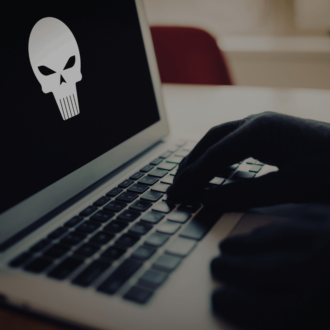 A imagem mostra uma mão enluvada digitando em um teclado de laptop, com uma tela exibindo um ícone de uma caveira estilizada. Este tipo de ícone frequentemente é associado a malware ou atividades cibernéticas maliciosas, sugerindo um cenário de ataque hacker.