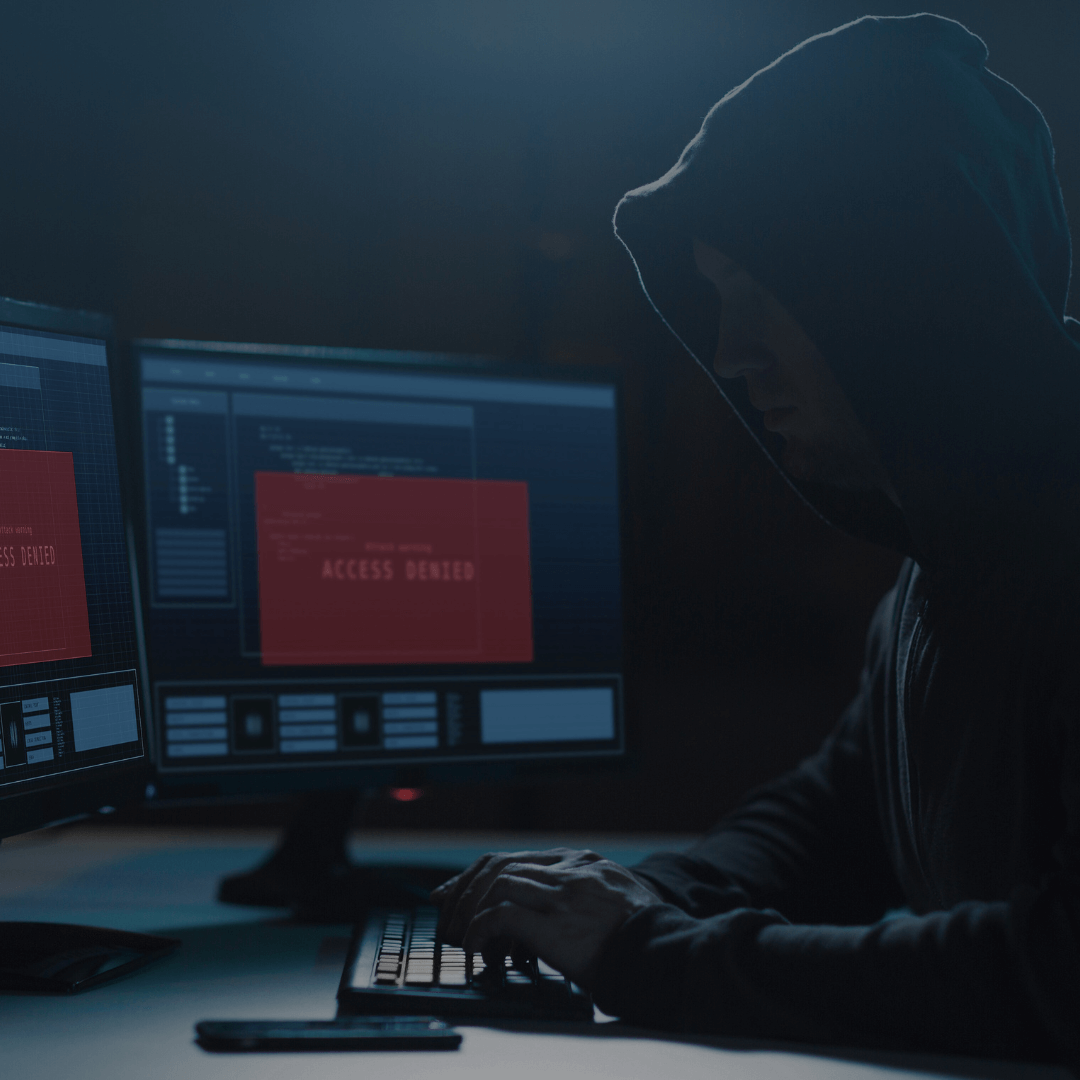 Imagem de uma pessoa com capuz sentada em frente a dois monitores, em um ambiente escuro. Nas telas, aparece uma mensagem de "Acesso Negado", sugerindo uma tentativa de invasão ou atividade cibernética maliciosa. A figura está digitando no teclado, o que reforça o cenário de cibersegurança e possível ataque hacker.