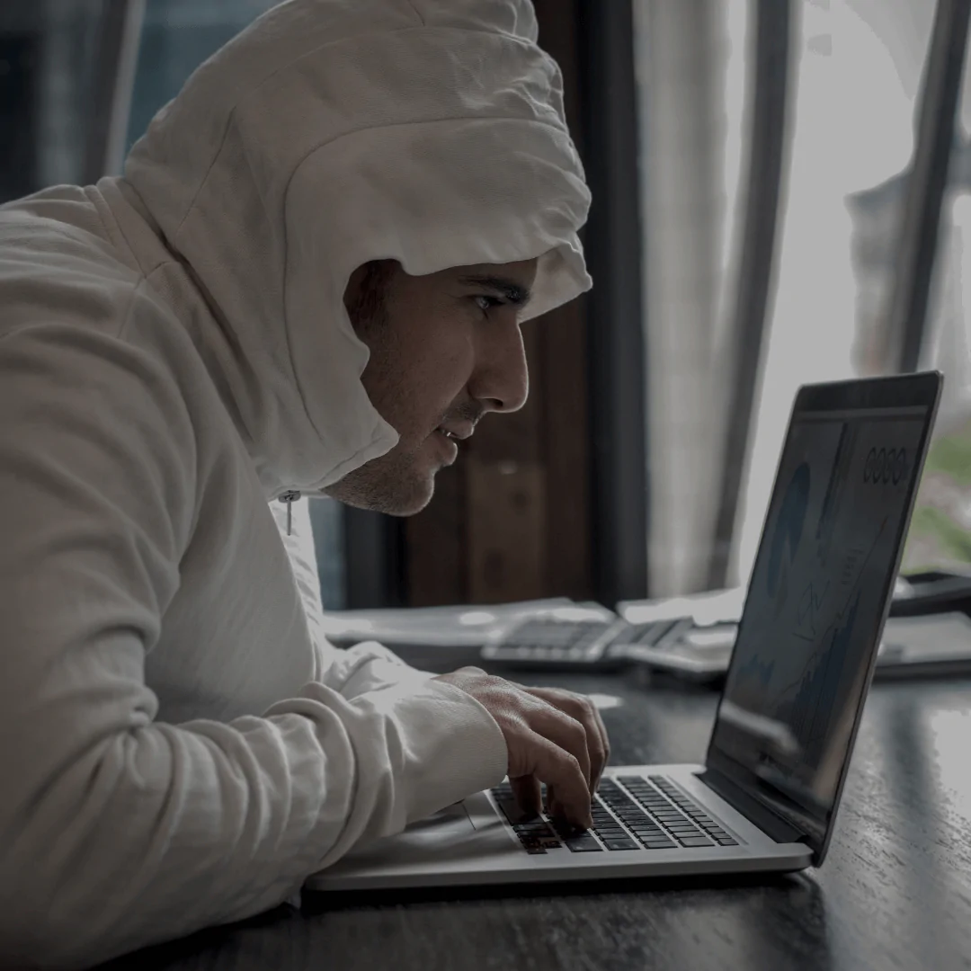 Homem vestindo um moletom com capuz branco, inclinado em frente a um laptop em um ambiente interno, aparentemente concentrado em algo na tela. A imagem transmite a ideia de uma atividade digital, como trabalho ou análise de dados, com um fundo ligeiramente desfocado.