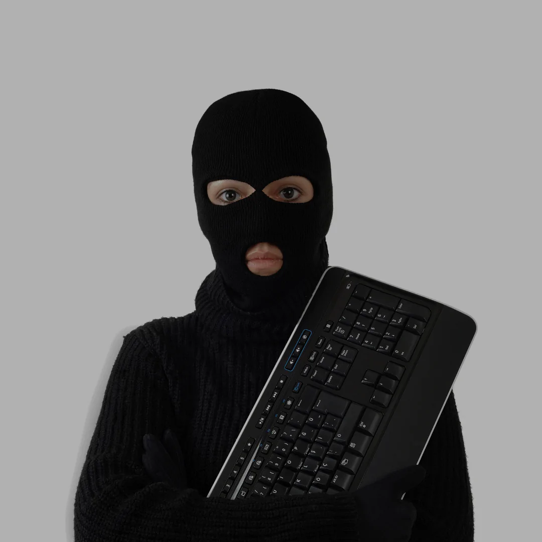 Pessoa usando balaclava preta e suéter escuro, segurando um teclado de computador. A imagem representa visualmente um conceito de cibercrime ou hacker.