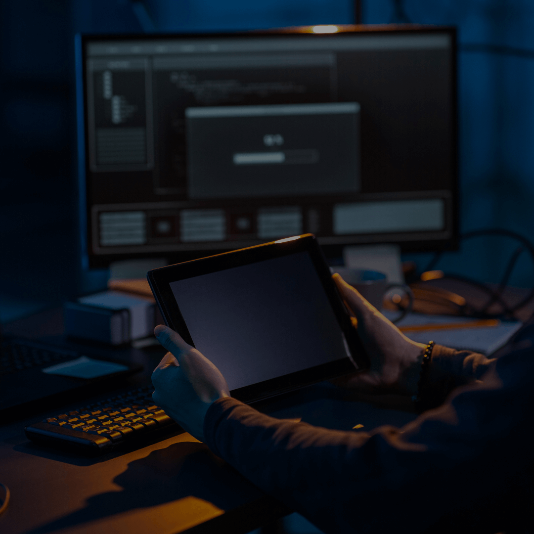 Pessoa segurando um tablet em um ambiente de escritório escuro, com uma tela de computador ao fundo mostrando uma interface de login. A cena sugere uma situação de trabalho em cibersegurança, destacando a proteção de dispositivos digitais e o acesso seguro a sistemas corporativos.