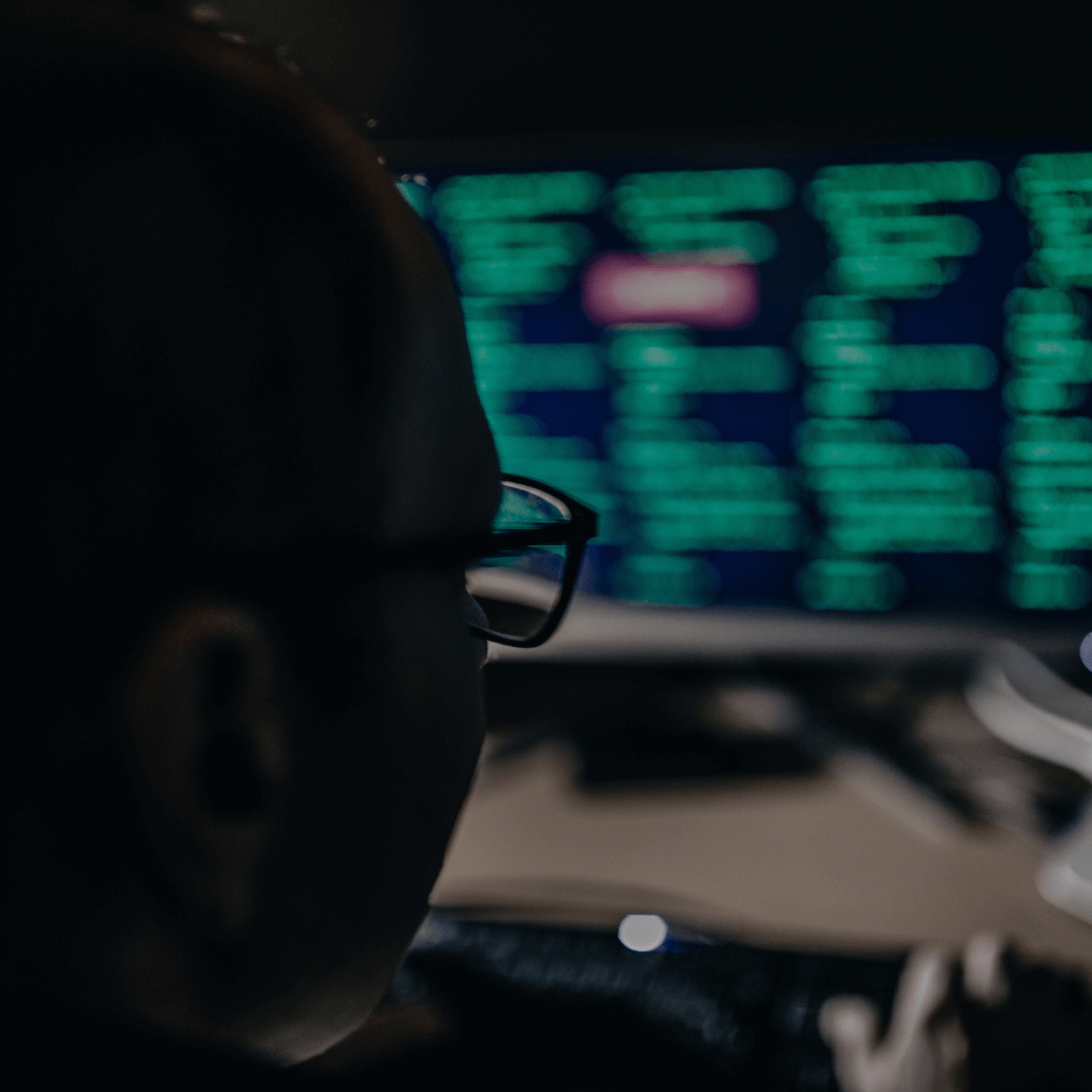 Pessoa observando uma tela de computador com linhas de código em verde, indicando um ambiente de trabalho em cibersegurança. A imagem está em baixa iluminação, com foco no monitor e nos reflexos dos códigos nos óculos do indivíduo, criando uma atmosfera de análise e vigilância digital.