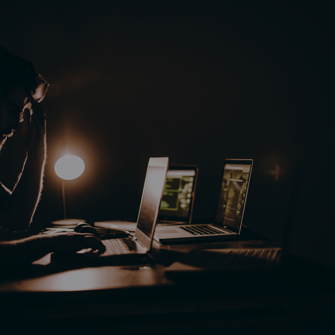 Pessoa trabalhando em uma sala escura, iluminada apenas por uma lâmpada e as telas de três laptops. A pessoa parece concentrada ou pensativa, com uma das mãos na cabeça, enquanto o ambiente sugere uma atmosfera de trabalho intenso, possivelmente em atividades de programação ou cibersegurança.