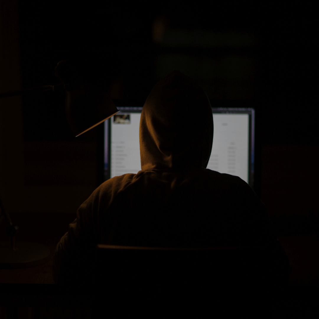 Pessoa de capuz sentada em frente a um computador em um ambiente escuro. A tela está parcialmente iluminada, revelando algumas informações indistintas, enquanto uma luminária direcionada para o monitor destaca o contorno da pessoa. A cena sugere um ambiente de trabalho reservado ou potencialmente relacionado a atividades de segurança digital.