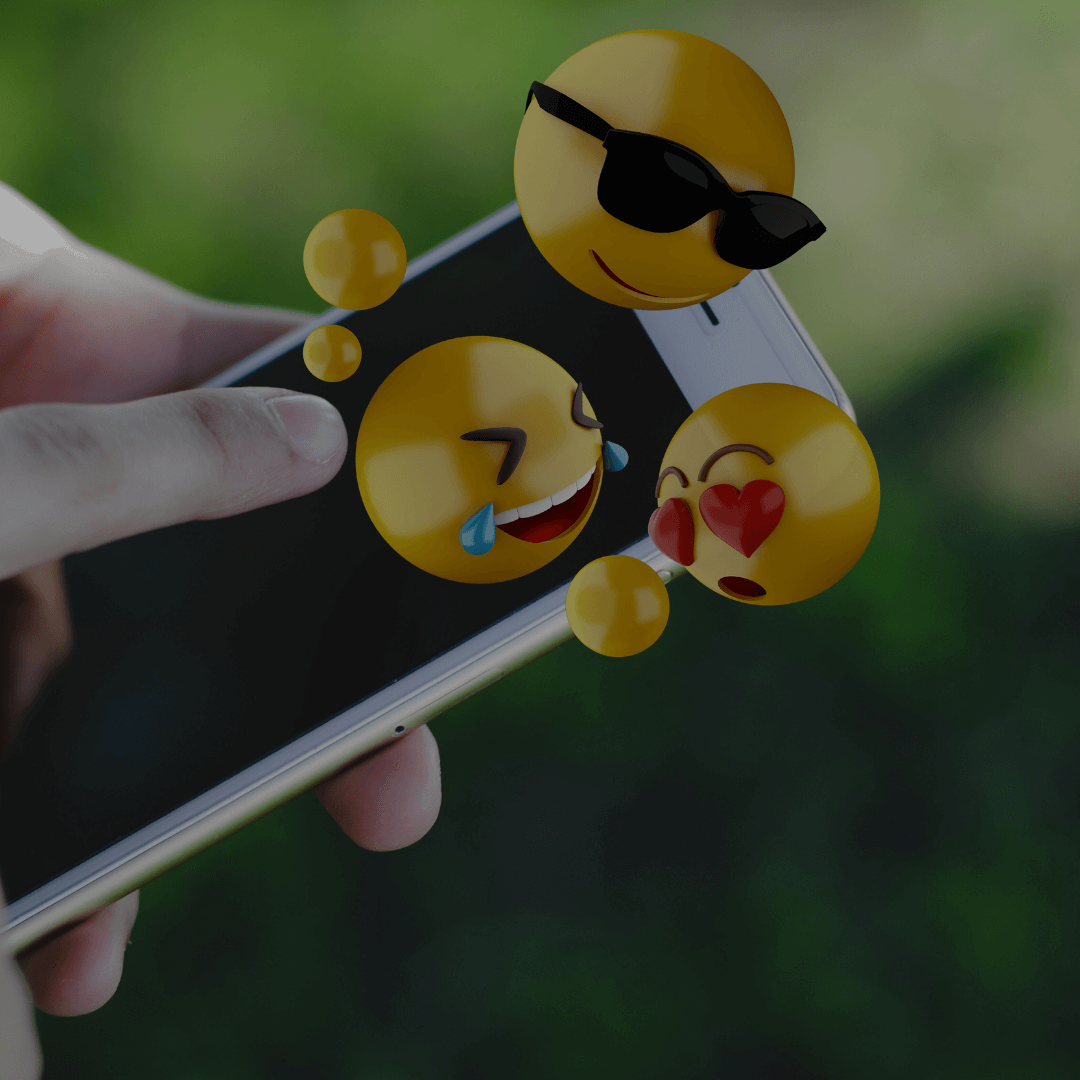 Uma mão segurando um smartphone de onde saem emojis em 3D flutuando. Os emojis representam emoções variadas, incluindo um emoji rindo com lágrimas, um emoji com óculos escuros e um emoji com olhos de coração, simbolizando diferentes sentimentos e reações que podem ser transmitidos digitalmente.