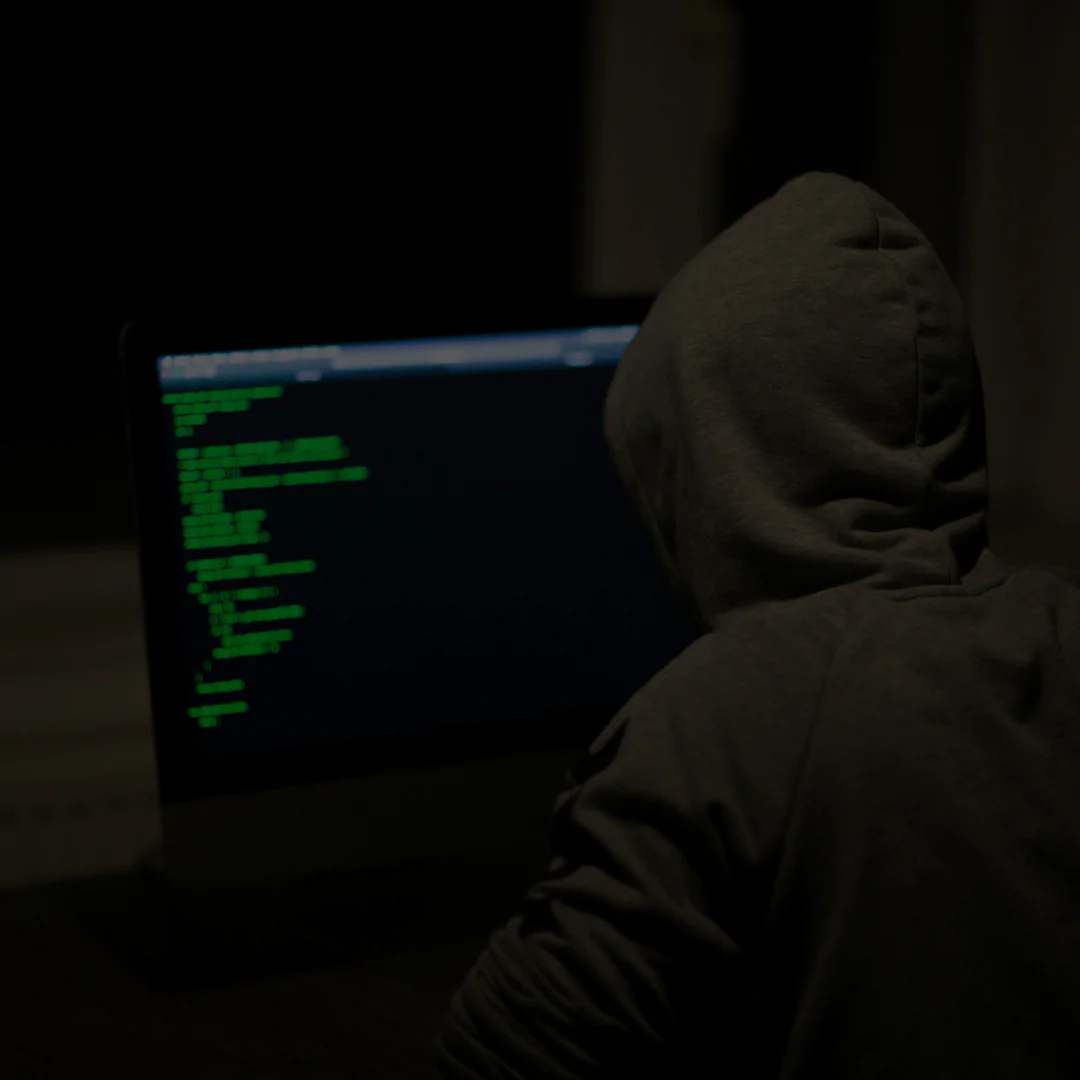 Uma pessoa vestindo um moletom com capuz está sentada em frente a um monitor de computador em um ambiente escuro. A tela exibe linhas de código ou comandos em texto verde, sugerindo atividades relacionadas a programação, segurança cibernética ou hacking. A iluminação mínima e o ângulo de trás destacam o tom misterioso da cena.