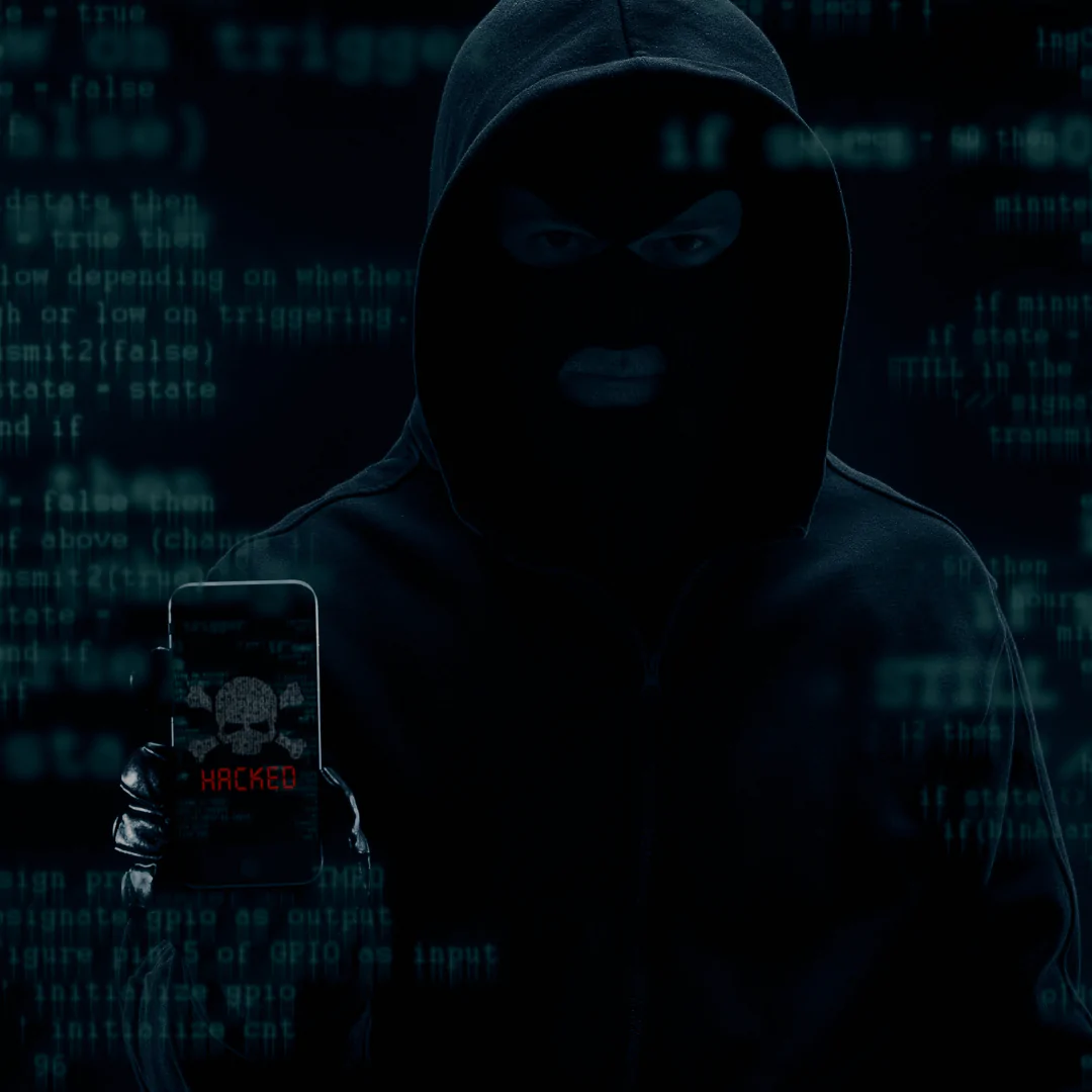 Homem vestido com capuz e máscara de ski preta, segurando um celular com uma imagem de um crânio e ossos cruzados na tela, acompanhada da palavra "HACKED" em vermelho. O fundo está repleto de códigos e texto digital em azul, representando um ambiente virtual ou cibernético, com um tom escuro e ameaçador.