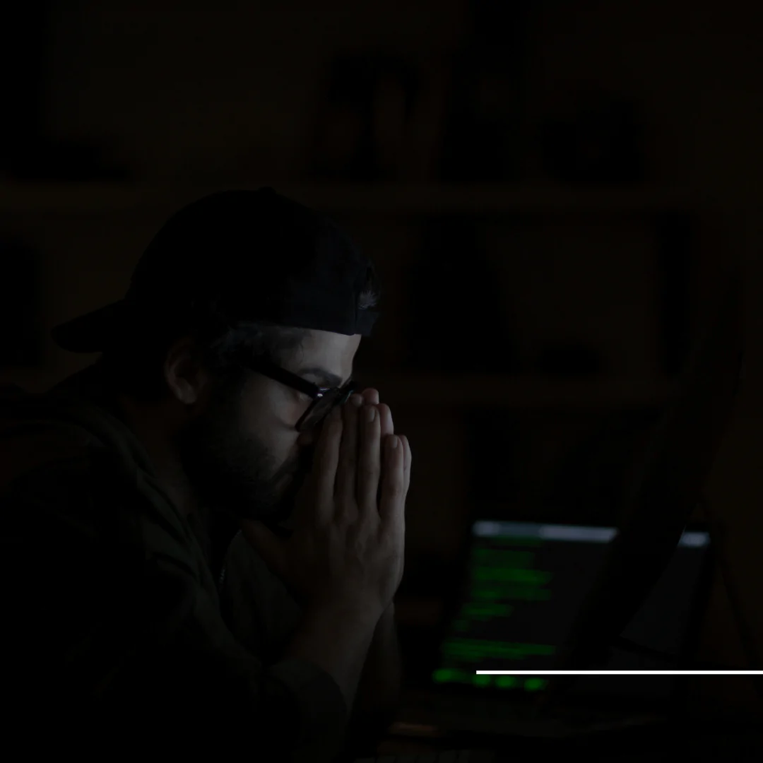 Um homem em um ambiente escuro, aparentando estar pensativo ou preocupado, com as mãos juntas na frente do rosto. Ele usa óculos, um boné preto e está olhando para a tela de um laptop, que exibe linhas de código verde. O ambiente está pouco iluminado, com destaque apenas para o rosto e a luz da tela do computador.