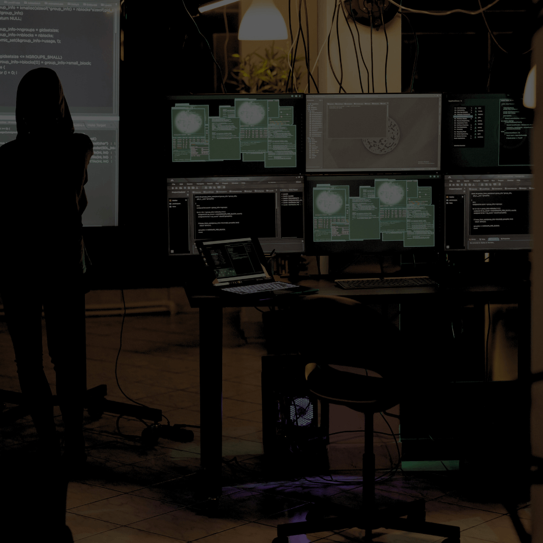 Uma sala de monitoramento cibernético com múltiplos monitores exibindo interfaces de software de segurança, incluindo gráficos e linhas de código. Ao fundo, uma pessoa em pé, parcialmente na sombra, observando as telas. O ambiente possui iluminação baixa e cabos visíveis conectando os equipamentos, reforçando o aspecto técnico e operacional do espaço.