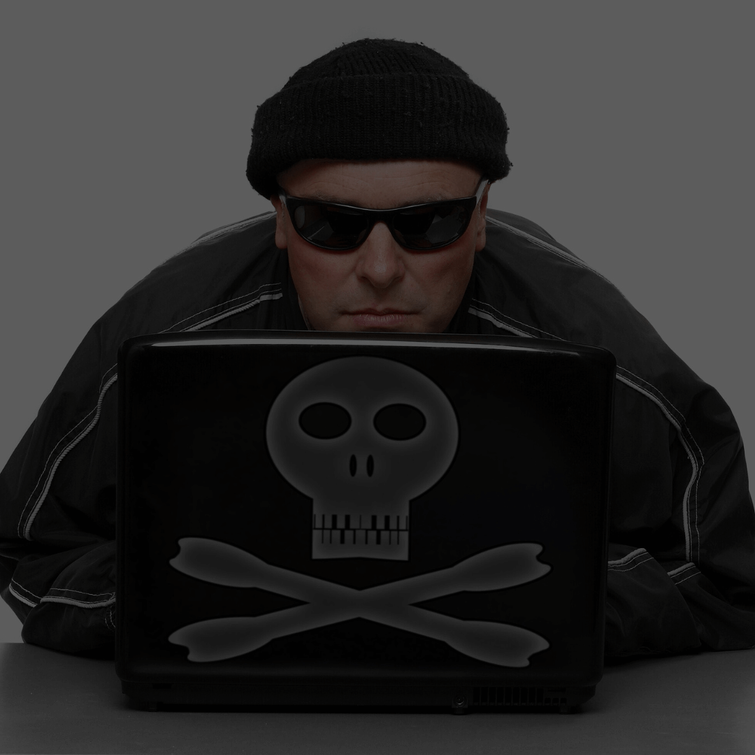 Homem usando um gorro preto e óculos escuros, olhando para a tela de um laptop. O laptop tem um adesivo com uma caveira e ossos cruzados, simbolizando atividades relacionadas a hackers ou cibersegurança. O fundo da imagem está escurecido, destacando o homem e o dispositivo.